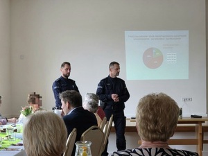 Dwóch policjantów, przed nimi osoby starsze siedzące przy stolikach. W tle wyświetlana prezentacja.