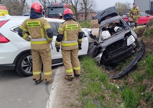 Miejsce wypadku drogowego. Widać dwa rozbite pojazdy i trzech strażaków.