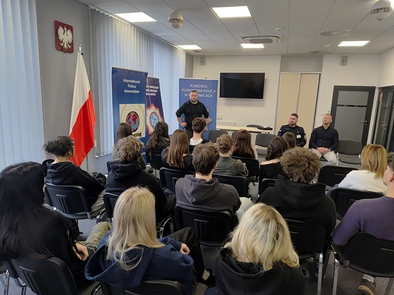 Policjant w stroju cywilnym, a przed nim uczniowie siedzący na krzesłach.