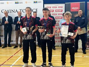 Dwóch chłopaków i dziewczyna z medalami i nagrodami. W tle policjanci i inne osoby.