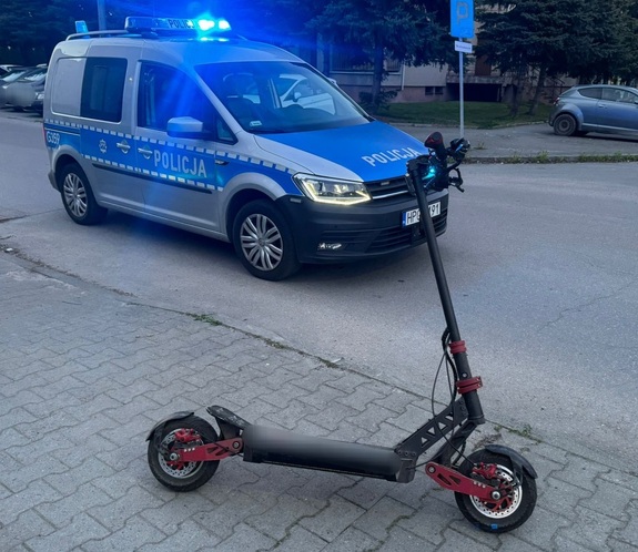 Na zdjęciu oznakowany radiowóz i hulajnoga elektryczna. W tle blok i samochody.