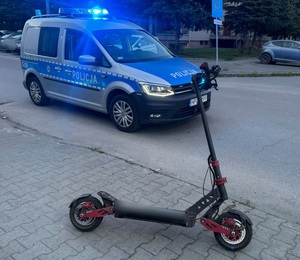 Na zdjęciu oznakowany radiowóz i hulajnoga elektryczna. W tle blok i samochody.
