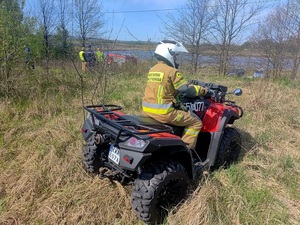 Strażak na quadzie. W tle radiowóz, wóz strażacki oraz policjanci i strażacy.