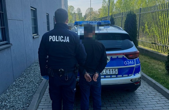 Na tle radiowozu policjant trzymający zatrzymanego mężczyznę, który ma założone kajdanki na ręce trzymane z tyłu