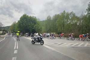 Na zdjęciu widać kolarzy  oraz mężczyznę w odblaskowej kamizelce a także motocykl