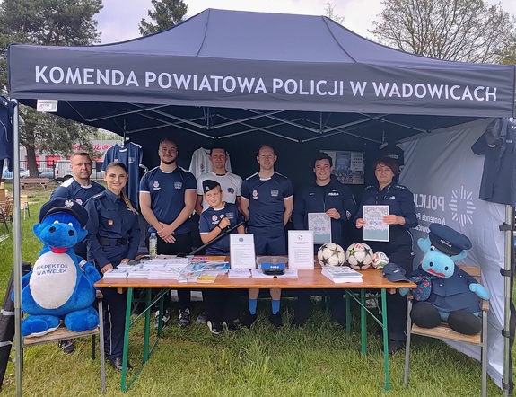 W namiocie czterech mężczyzn i chłopiec w sportowych strojach, policjant oraz dwie policjantki. Na stoliku gadżety.