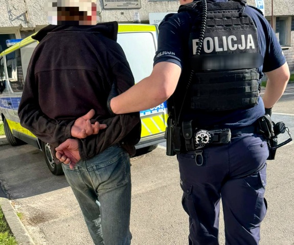 Umundurowany policjant prowadzący zatrzymanego mężczyznę. W tle radiowozów
