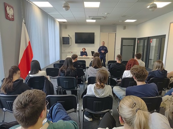 Policjantka siedząca przy stoliku, obok stoi policjant, a przed nimi młodzież siedząca na krzesłach