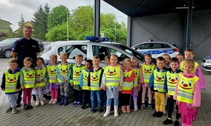 Na tle radiowozów przedszkolaki w odblaskowych kamizelkach, po lewej policjant