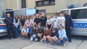Na tle radiowozu grupa osób, chłopców i dziewcząt. Po prawej i lewej stronie umundurowany policjant