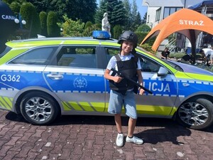 młoda dziewczynka ubrana w policyjne wyposażenie hełm, kamizelke w ręku trzyma pałkę typu tonfa, w tle radiowóz