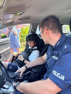 policjant w radiowozie obok siedzi chłopiec ubrany w sprzęt policyjny.