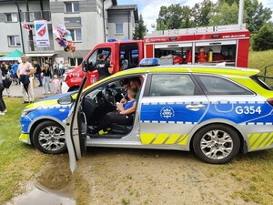 w radiowozie policyjnym mały chłopiec z dorosłym siedzi za kierownicą obok policjant, obok przechodzą uczestnicy festynu