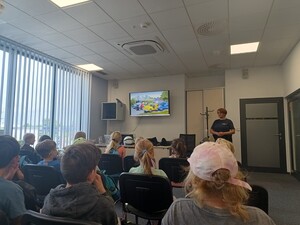 dzieci w sali sztabowej słuchają policjantki, w tle włączony film animowany