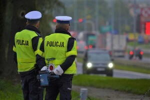Policjanci ruchu drogowego w odblaskowych kamizelkach. Jeden z nich trzyma w ręku miernik prędkości. W tle samochody.