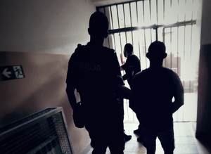Na zdjęciu widać postury dwóch policjantów oraz zatrzymanego mężczyzny na tle krat