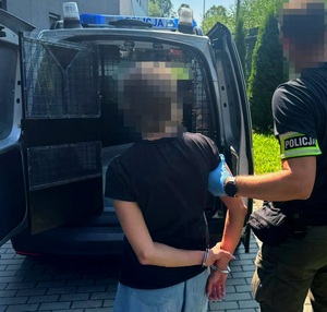 Nieumundurowany policjant, który ma na ręce założoną opaskę z napisem Policja trzyma kobietę. W tle radiowóz