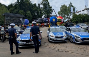 Na zdjęciu trzy radiowozy oraz dwóch policjantów stojących tyłem. W tle kibice.