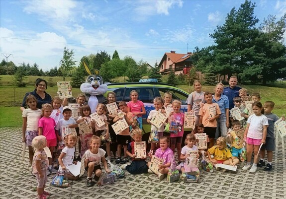 Na tle radiowozu grupa dzieci z dyplomami, dwie kobiety w stroju cywilnym, policjant i maskotka królika