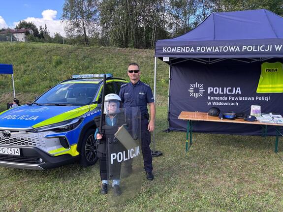 Na tle radiowozu i namiotu policjant i chłopiec ubrany w elementy policyjnego umundurowania.