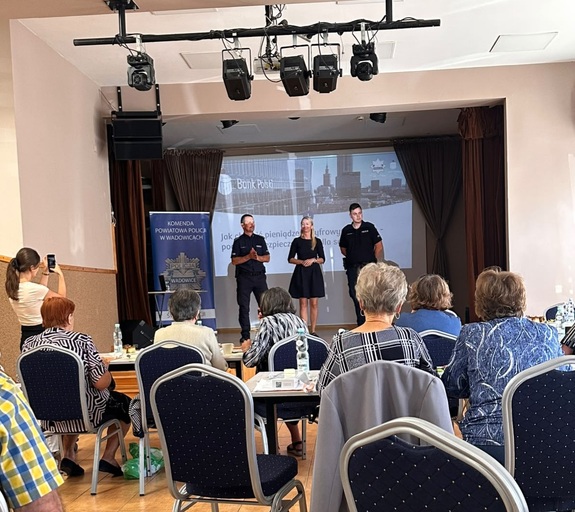 Na scenie dwóch policjantów i kobieta w stroju cywilnym. Przed nimi seniorzy siedzą na krzesłach