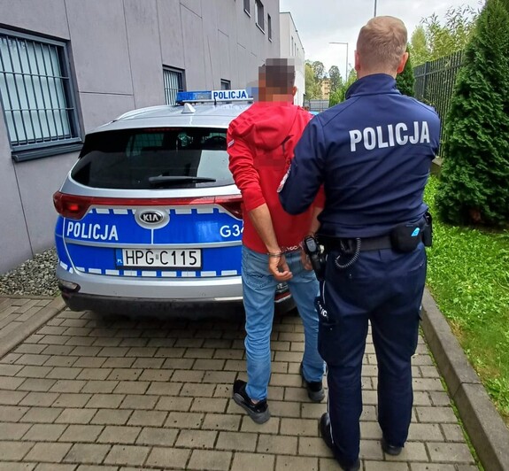 Na tle radiowozu umundurowany policjant, który trzyma zatrzymanego mężczyznę