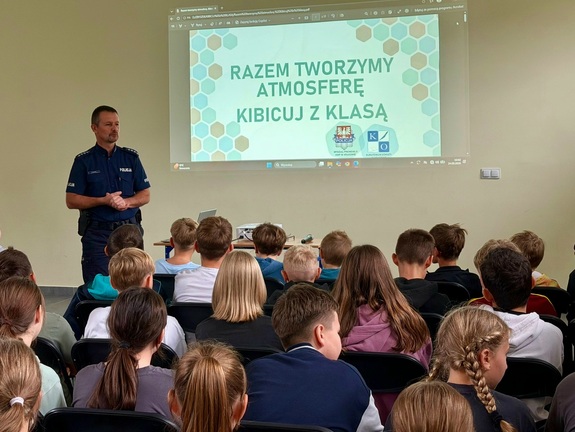 Na zdjęciu policjant, który wyświetla na ekranie prezentację, przed nimi siedzą dzieci