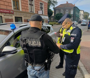 Umundurowany policjant i policjant w stroju cywilnym w kamizelce z napisem Policja. W tle budynki i samochody