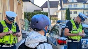 Na tle budynków i radiowozu dwóch policjantów ruchu drogowego i chłopak w kasku ochronnym