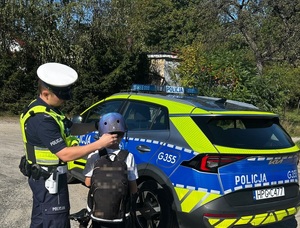 Policjant poprawiający kask chłopcu na rowerze. Obok radiowóz
