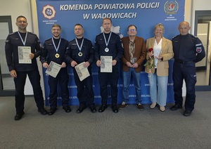 Czterech policjantów z medalami i dyplomami, a po prawej mężczyzna i kobieta w stroju cywilnym i umundurowany policjant