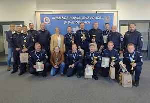 Na tle policyjnego baneru piętnastu policjantów, dwóch mężczyzn i kobieta w stroju cywilnym .