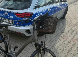 Na tle oznakowanego radiowozy rower z koszykiem na kierownicy