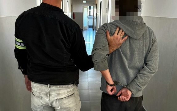 Po lewej stronie nieumundurowany policjant, który na lewej ręce ma założoną opaskę  z napisem Policja. Trzyma mężczyznę, który ma założone kajdanki na rekach trzymanych z tyłu