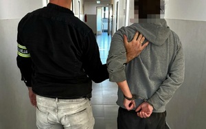 Po lewej stronie nieumundurowany policjant, który na lewej ręce ma założoną opaskę  z napisem Policja. Trzyma mężczyznę, który ma założone kajdanki na rekach trzymanych z tyłu