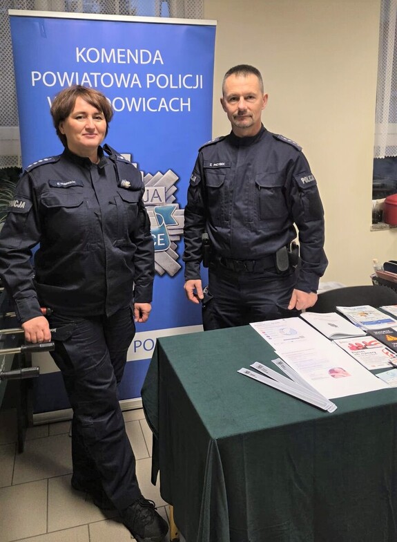 Na tle policyjnego baneru umundurowana policjantka i policjant. Na stoliku ulotki