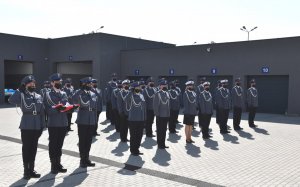 Policjanci i policjantki w umundurowaniu galowym stojący w kilku szeregach na placu komendy. Na zdjęciu po lewej stronie obok funkcjonariuszy stoi poczet flagowy, trzech policjantów. Funkcjonariusz stojący w środku trzyma w rękach złożoną flagę Polski.