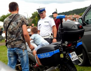 Umundurowany policjant ruchu drogowego stojący obok motocykla, na którym siedzi mały chłopiec. Po lewej stronie stoi tyłem mężczyzna w stroju cywilnym. W tle widać ludzi.