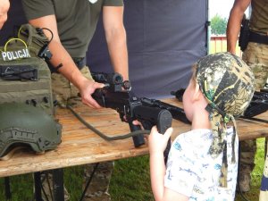 Mały chłopiec stojący tyłem trzymający w ręku pistolet. Po lewej stronie kamizelka kuloodporna. W tle umundurowani policjanci.