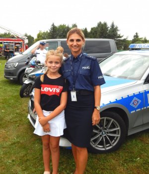 Umundurowana policjantka stojąca obok małej dziewczynki na tle radiowozu. W tle policjant ruchu drogowego, motocykl i samochód.