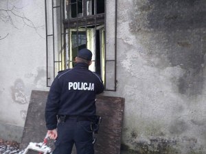 Umundurowany policjant stojący tyłem, sprawdzający przez okratowane okno czy wewnątrz znajdują się jakieś osoby