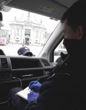 Umundurowany policjant siedzący w radiowozie, piszący w notatniku służbowym. W tle spacerujący ludzie oraz kościół.