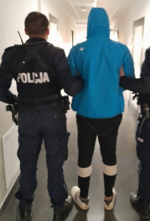Zatrzymany mężczyzna stojący tyłem. Ubrany w bluzę koloru niebieskiego z kapturem założonym na głowę i spodnie koloru ciemnego. Po jego lewej stronie stoi umundurowany policjant tyłem.