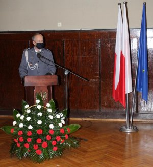 Policjant w umundurowaniu galowym z założoną na twarzy maseczką, stojący przed mównicą, przed którą znajduje się kosz z kwiatami biało-czerwonymi oraz mikrofon. Obok wiszą dwie flagi Polski oraz flaga Unii Europejskiej.