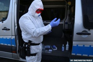 Policjant ubrany w biały kombinezon ochronny, na dłoniach założone ma niebieskie ochronne rękawiczki, stojący na tle radiowozu