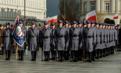 Policjanci w umundurowaniu galowym, stojący na placu w szeregach, trzymający flagi Polski. W tle widać budynki