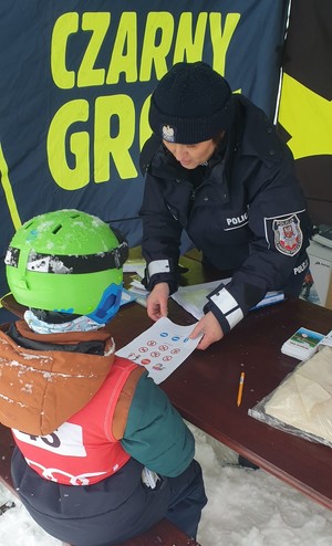 Umundurowana policjantka pokazująca na kartce znaki drogowe dziecku ubranemu w strój narciarski i kask. Dziecko siedzi tyłem. W tle napis Czarny Groń.