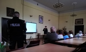 Na zdjęciu widać kilku seniorów siedzących na krzesłach, przed nimi umundurowana policjantka, po lewej stronie umundurowany policjant tyłem.