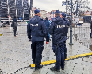 Trzech umundurowanych policjantów stojących tyłem. W tle fragment sceny oraz publiczność.