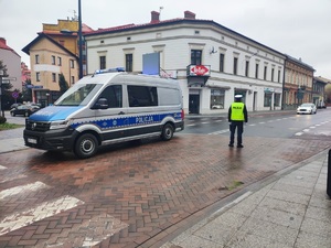 Na zdjęciu widać policyjny radiowóz obok umundurowanego policjanta ruchu drogowego, w kamizelce odblaskowej z napisem Policja. W tle budynki i jadący samochód koloru białego.